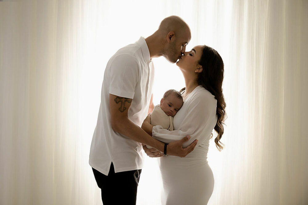 A family portrait of a man and women facing each other and kissing both holding their newborn between them against a light and bright backdrop in a professional photography session in Southampton, New Jersey.