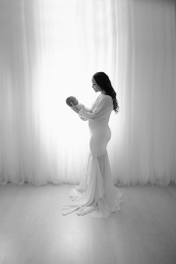 Black and white portrait of a woman standing to the side and holding her newborn on her tummy looking at her for her newborn photography photos in Westampton, New Jersey.