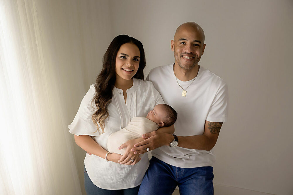 A man and a woman standing next to each other and smiling as she holds their new baby in her arms taking during their professional baby photography session in Medford, New Jersey.