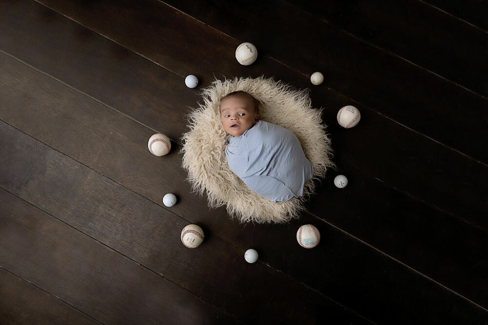 A baby resting on fuzzy blanket surrounded by baseballs and golf balls awake for a dream in-studio newborn session in Tabernacle, New Jersey.
