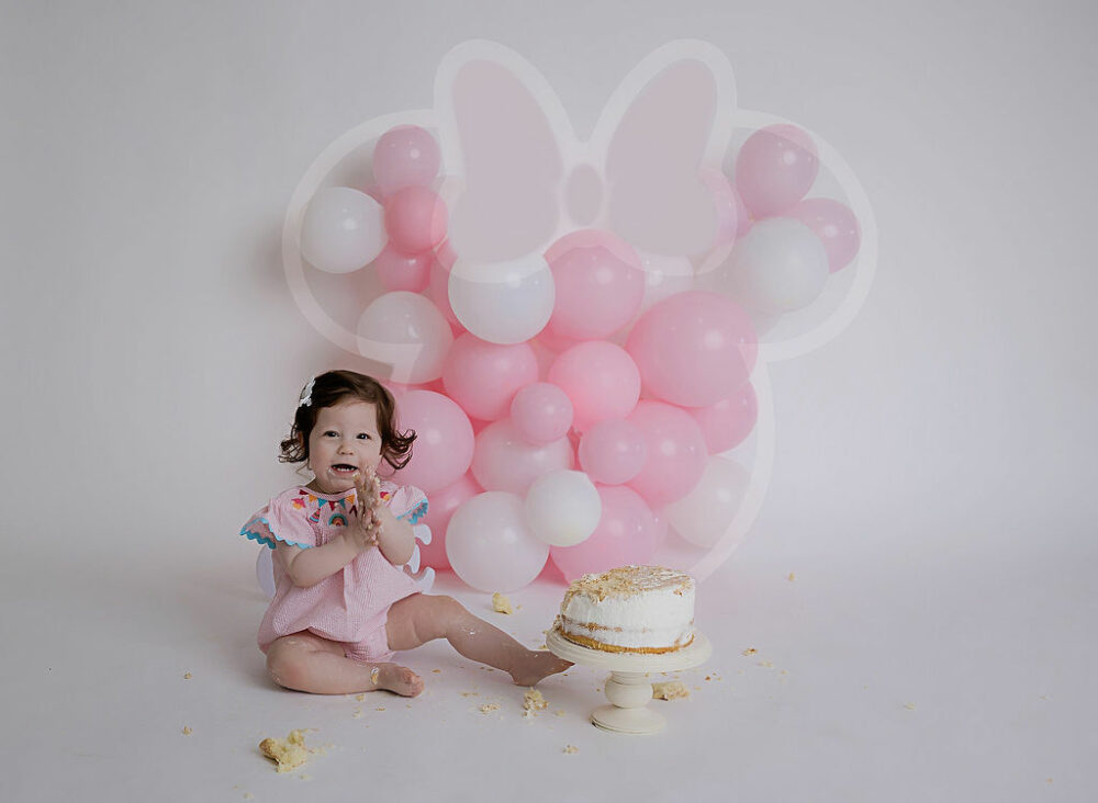 A toddler girl sitting beside a cake and having fun during her pink mini mouse -themed session in Deptford, New Jersey.