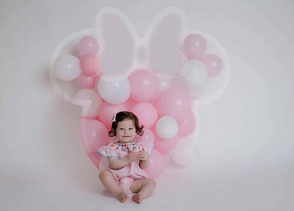 A toddler girl sitting and smiling for her baby portraits taken during her pink mini mouse first birthday session in Medford, New Jersey.