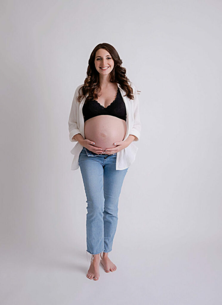 A woman holding her belly and wearing jeans and open blouse showing her tummy for the best maternity photography in Southampton, New Jersey.