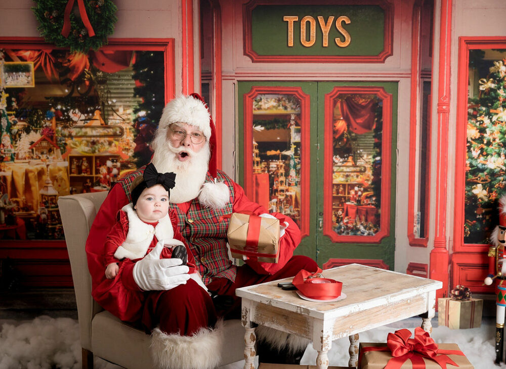 A infant girl posing with Santa during her south Jersey Christmas Santa mini session in toy shop set in Moorestown, New Jersey.