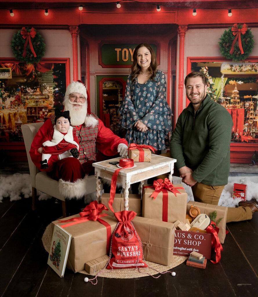 A family portrait of a couple posing with Santa Claus and his toy shop with a newborn for South Jersey Christmas Santa mini session in Pemberton, New Jersey.