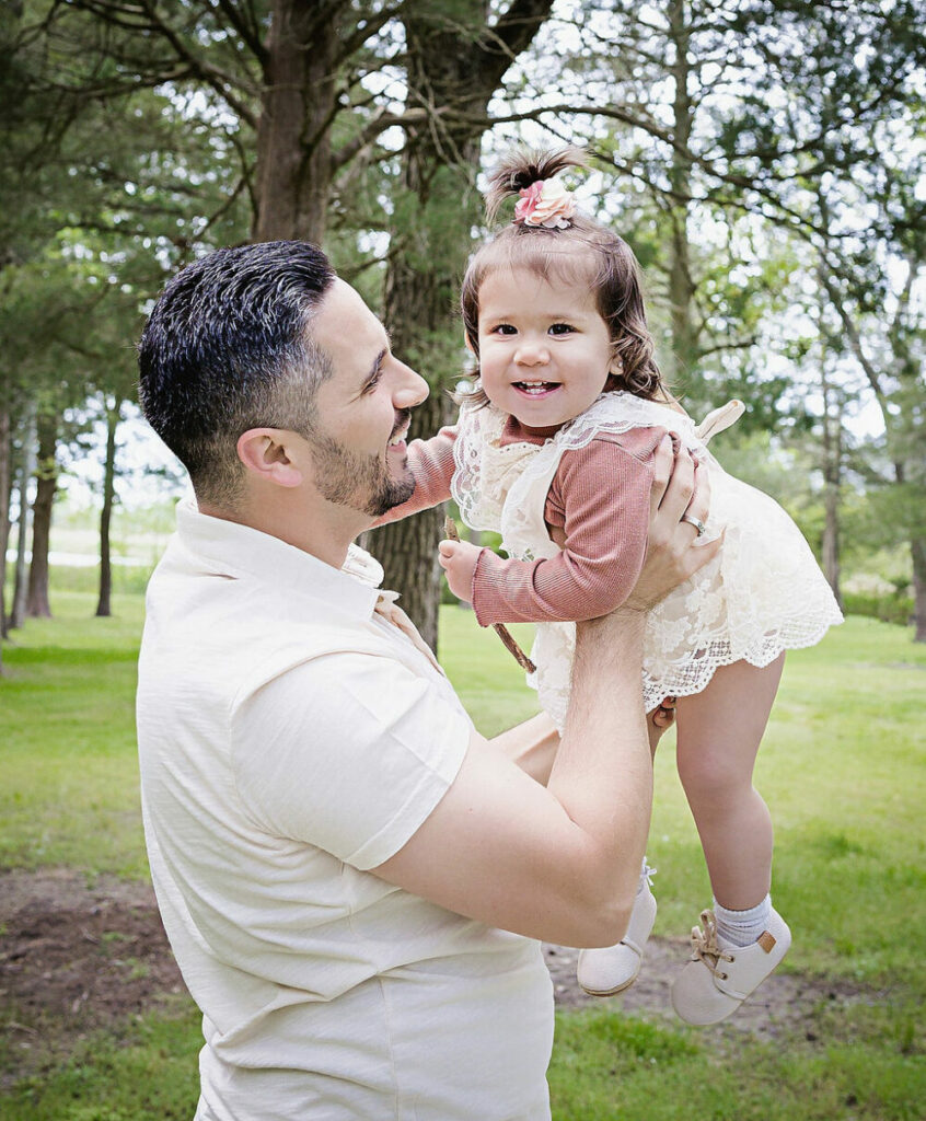A father-and-daughter picture of a man with his toddler girl. He is picking her up while she smiles and looks at the camera for their spring outdoor family session in Westampton, New Jersey.