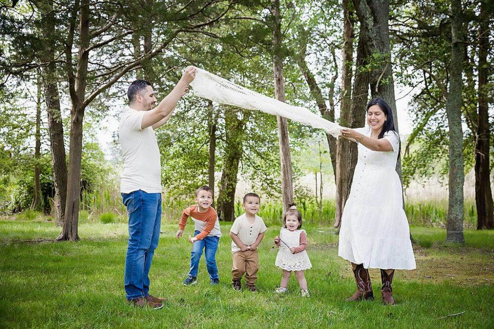 A lifestyle photo of a man and woman raising a blanket in the air as their kids stand underneath as they play during their spring outdoor family session in Cherry Hill, New Jersey.