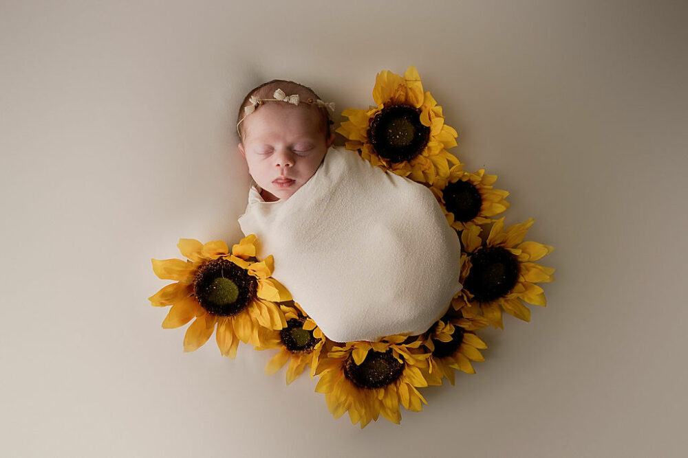 a baby girl sleeping on her back and swaddled in a wrap, adorned with flowers for her sunflower newborn session in Southampton, New Jersey.