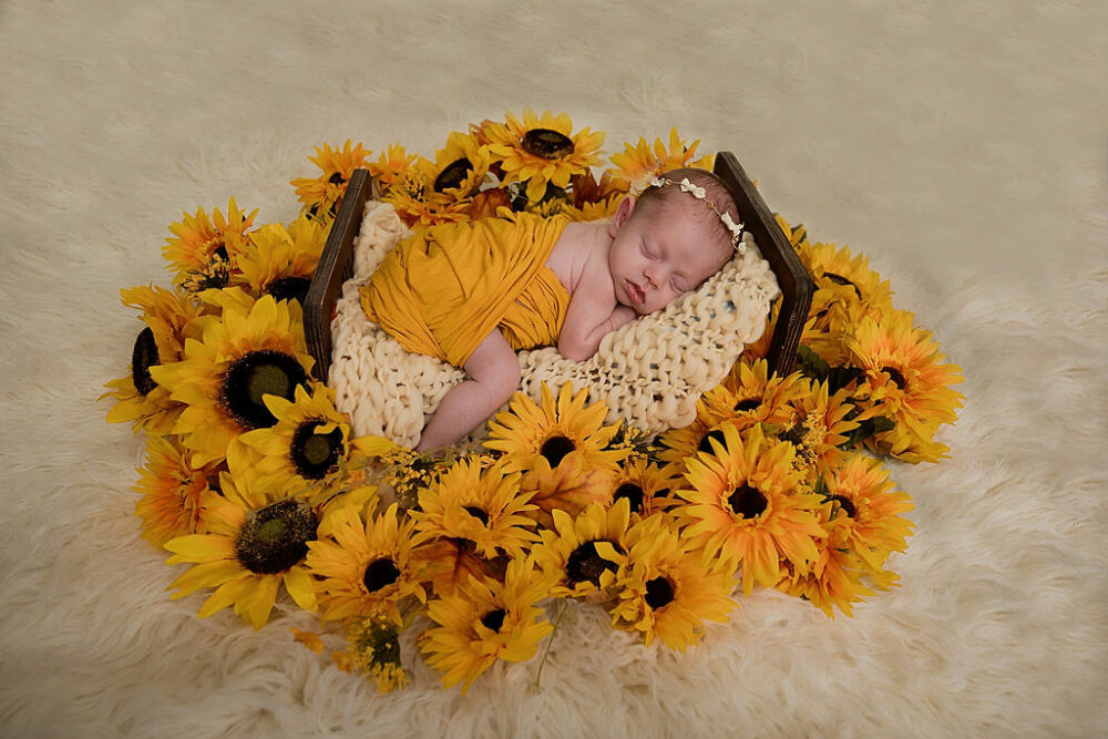 An in fact sleeping on her belly, wrapped and resting on a textured blanket, on a crib photography prop adorned with flowers for her sunflower newborn session in Deptford, New Jersey.
