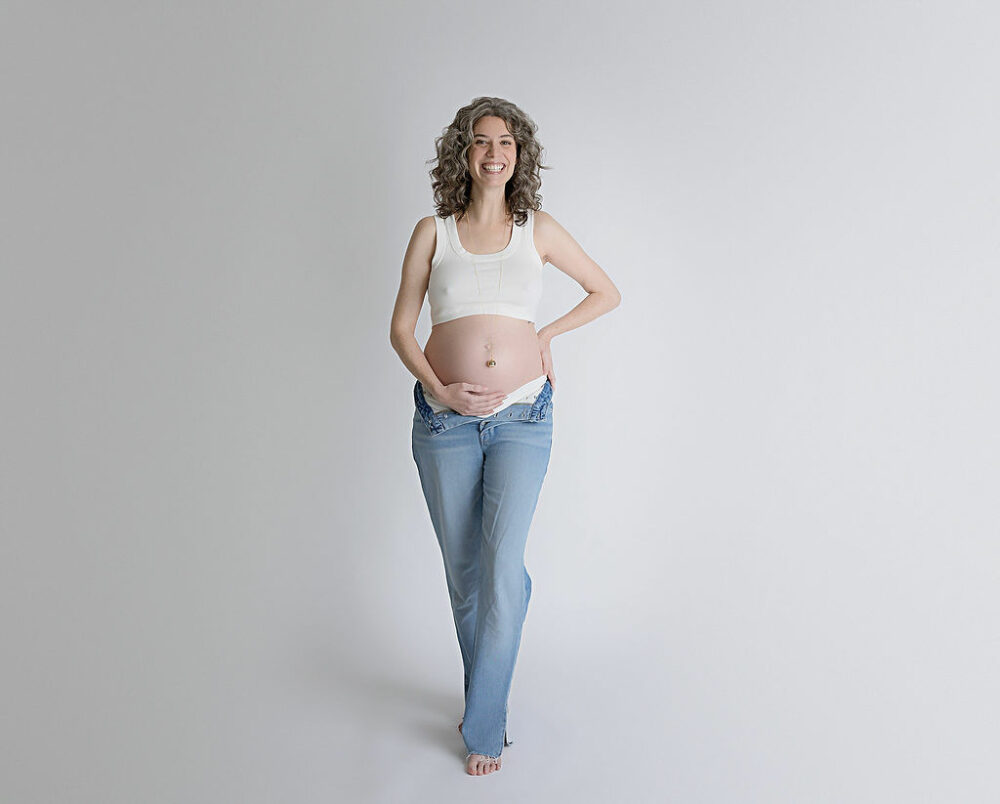 A woman standing and smiling as she hold her belly and is lightly posed for her maternity session in Tabernacle, New Jersey.
