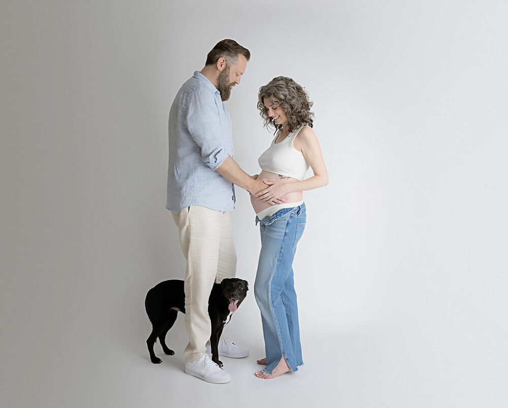 A man and a woman facing each other as they both told her that he wearing linen outfits and posing with their dog for a family photo during her maternity session in Crosswicks, New Jersey.