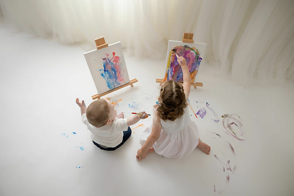 A sibling portrait of a boy and a girl sitting on the floor painting on canvas on easels for their colorful milestone session in Southampton, New Jersey.