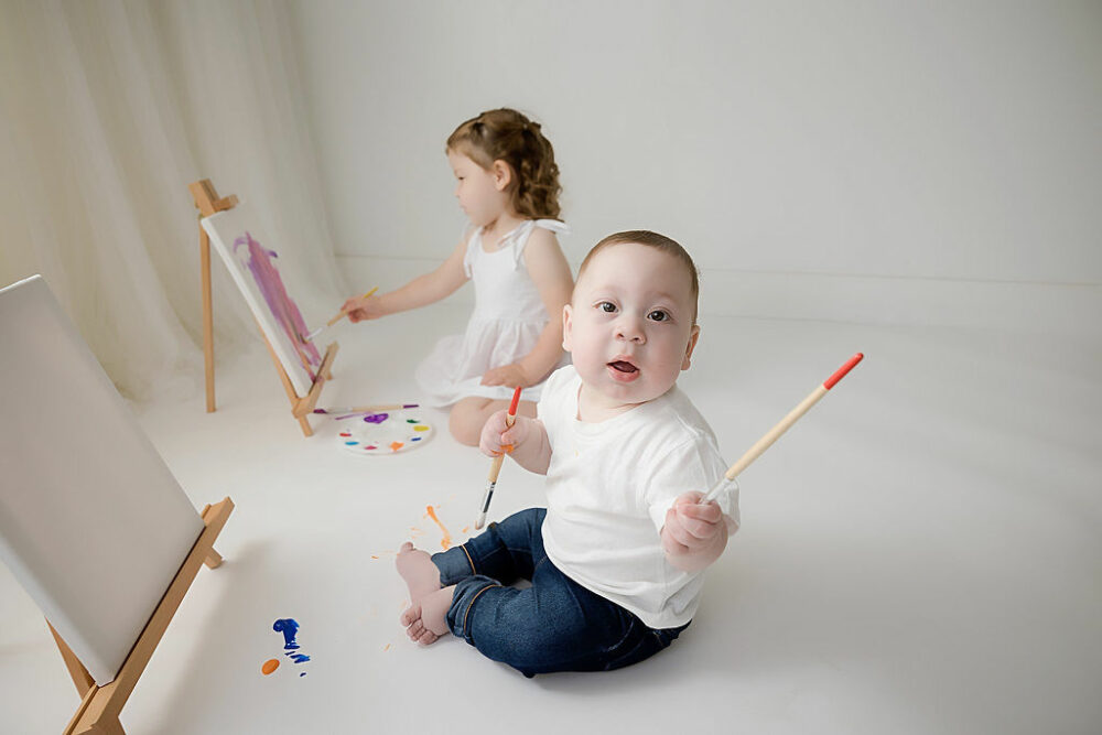 A infant boy facing camera as his sister is painting behind him for her colorful milestone session in Eastampton, New Jersey.