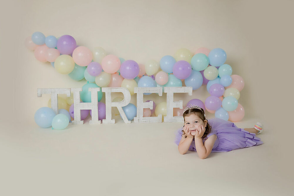A toddler girl resting on tummy with her head plopped on her hands with colorful balloon garland as a backdrop with large letters that spell the word three, posing for her colorful milestone session in Tabernacle, New Jersey.