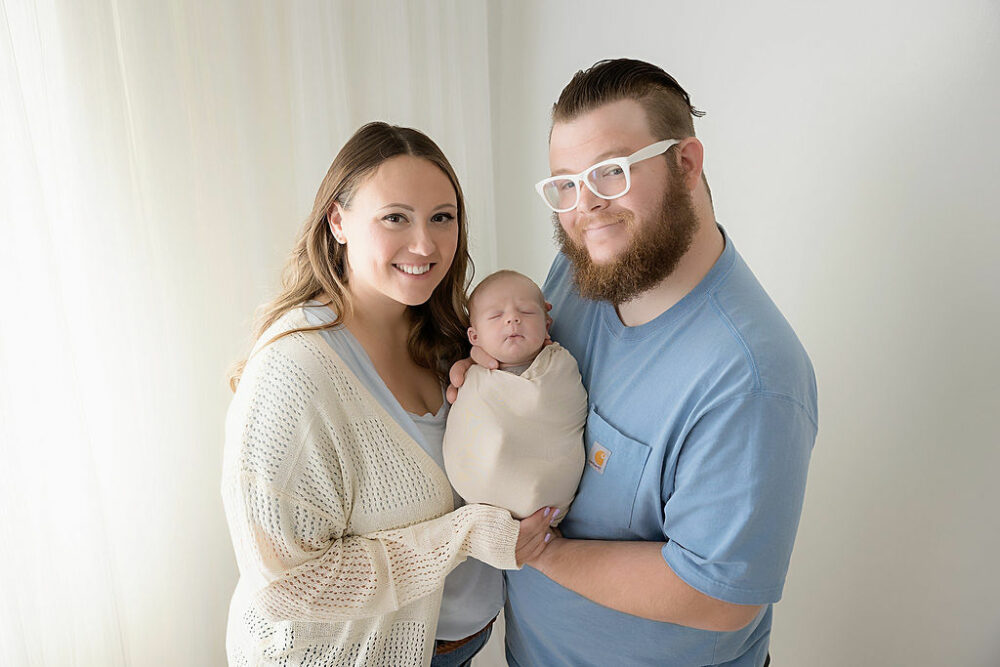 A newborn boy sleeping while being held by both of his parents who are smiling and posing wearing light colors against a light backdrop for their professional newborn and fall family session in Southampton, New Jersey.