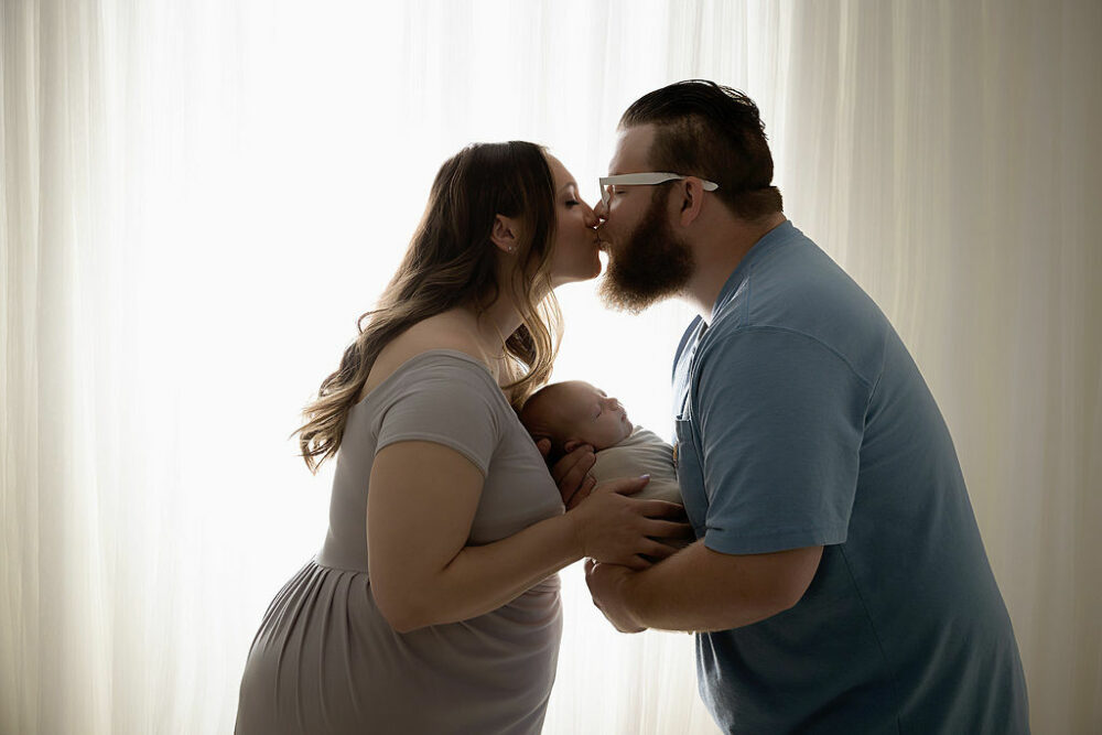 A man and a woman facing each other and kissing on the lips as they hold their newborn son between them against a light and bright backdrop for their infant photography session in Westampton, New Jersey.