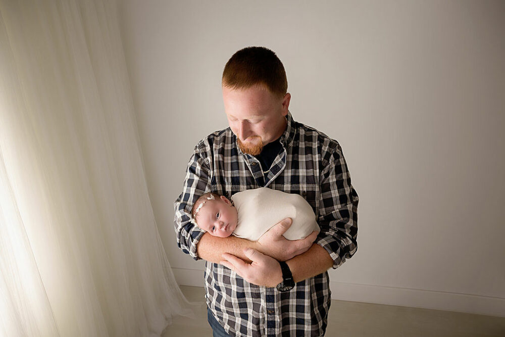 A man holding his newborn in his hands as she is wrapped and comfortable for a in-studio princess newborn session in Westampton, New Jersey.