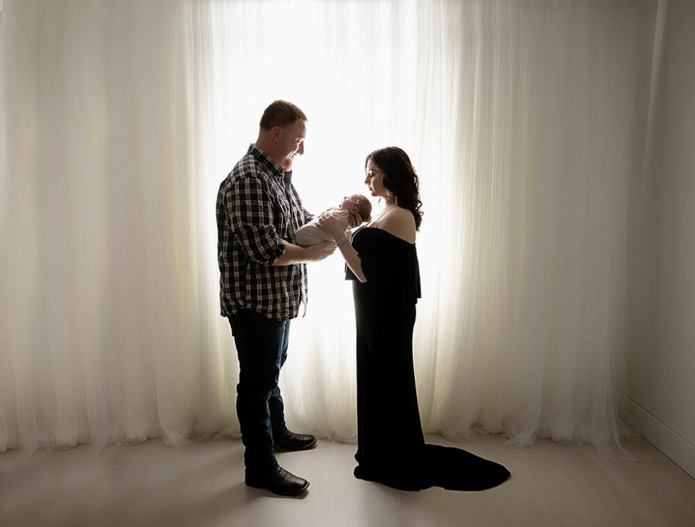 A man and a woman facing each other while holding their baby against a light and bright backdrop posing for an in-studio princess newborn session in Eastampton, New Jersey.
