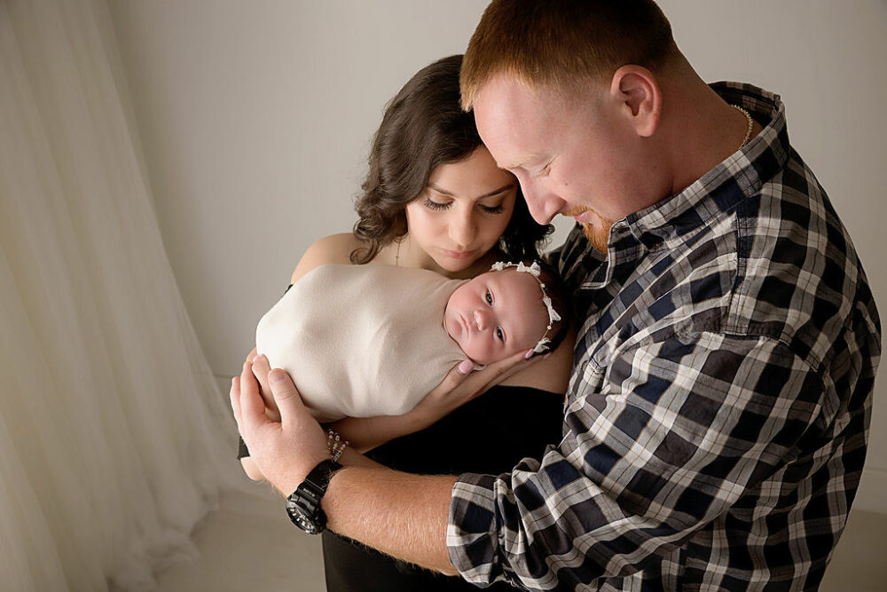 A close-up of a couple looking at their newborn girl as a both embrace her in their arms for their family portraits taken during her in studio princess newborn session in Medford, New Jersey.