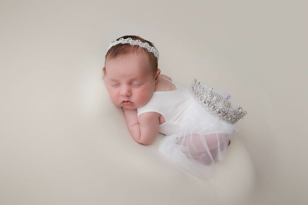 A newborn girl sleeping on her tummy with her knees tucked in wearing cute ballet outfit and sparkly headband for her in-studio princess newborn session in Cherry Hill, New Jersey.