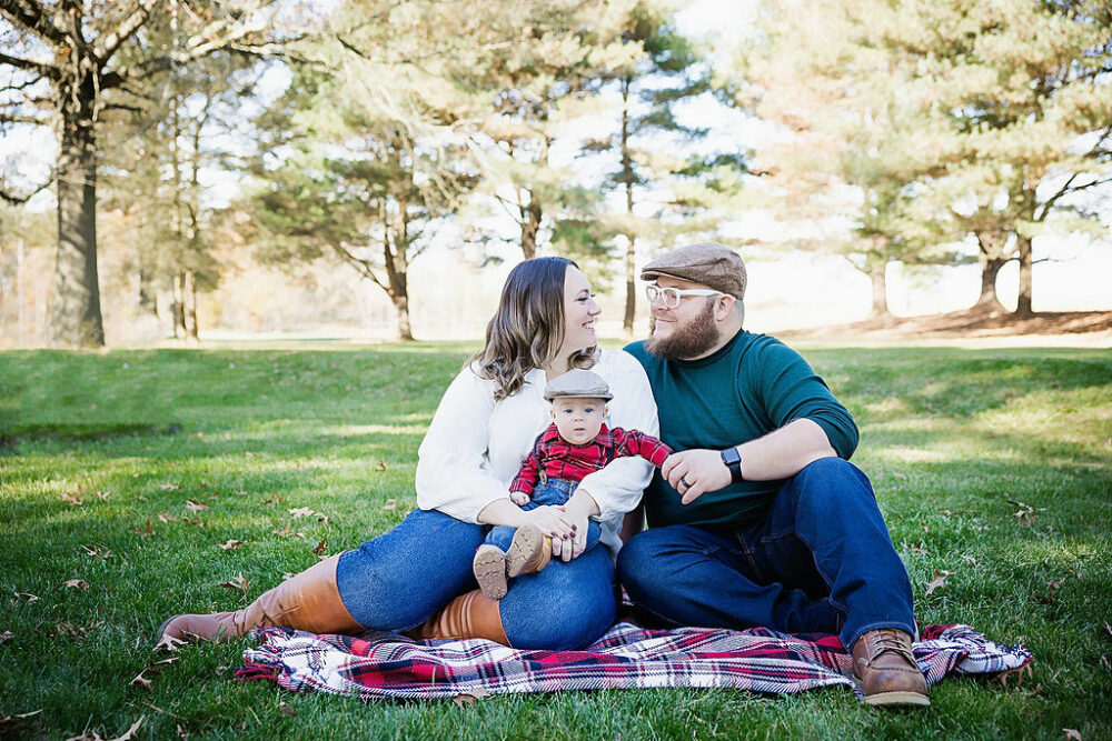 A family photo of a man and woman sitting on the ground facing each other and smiling as she holds her newborn son on her lap for a fall family photo shoot in Mount Laurel, New Jersey.