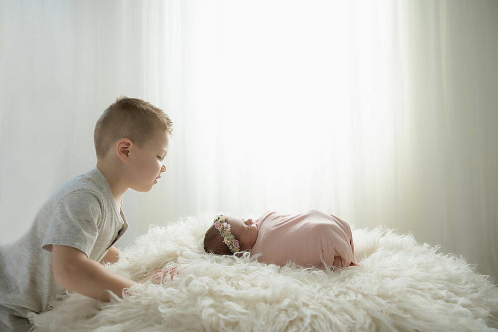 A sibling photo of a toddler boy looking down at his baby sister as she sleeps during her baby photography session in Eastampton, New Jersey.