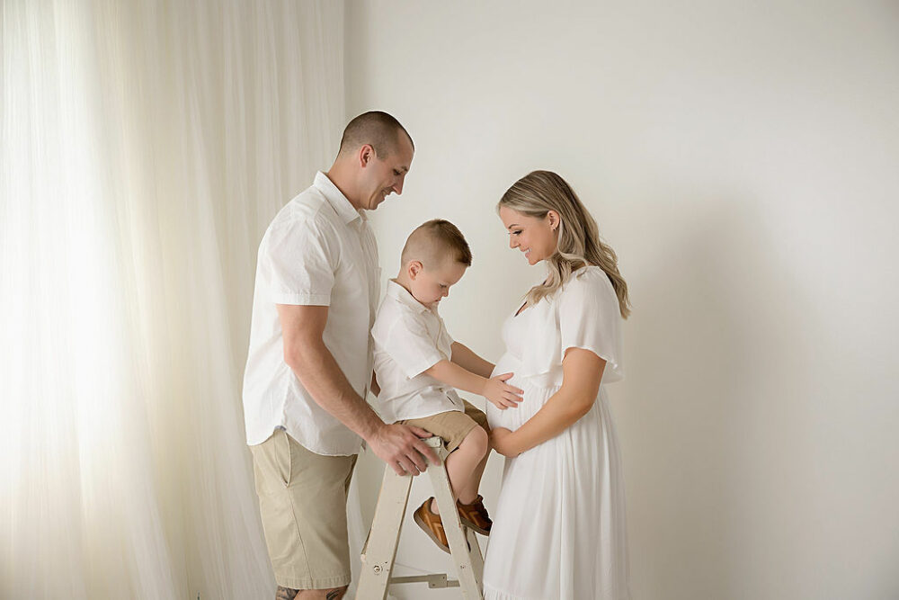 A family photo of a man and woman facing each other as a toddler boy sit in between them and hold her belly for their family maternity session in Moorestown, New Jersey.