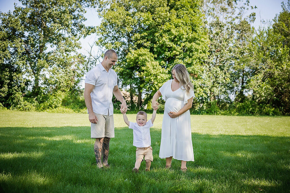 A lifestyle photo of a man and woman holding their toddler son's hands as they pose for outdoor maternity session in Tabernacle, New Jersey.