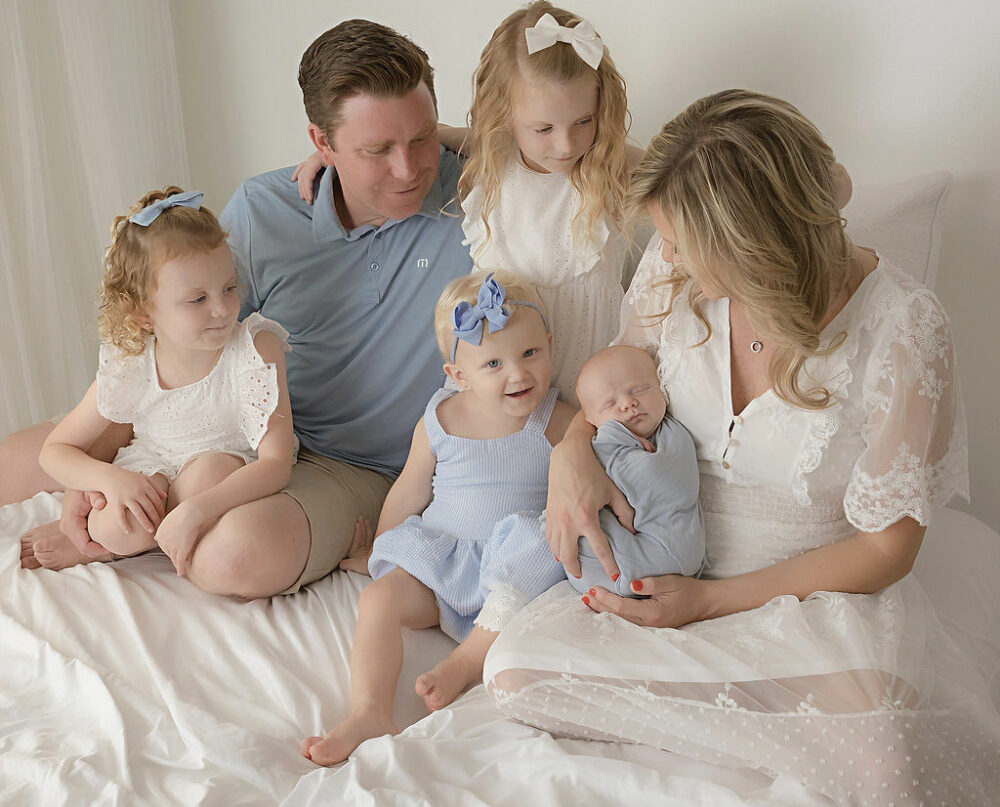 A man and woman and their four children sitting on bed photography set for their newborn photos taken in Southampton, New Jersey.