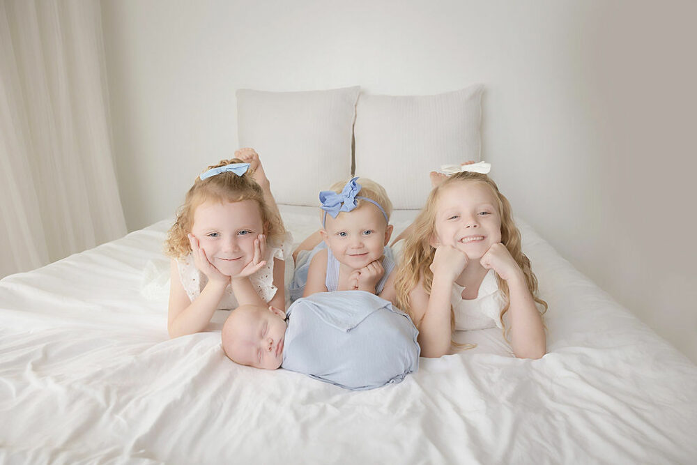 A family portrait of three sisters lightly posed while next to theirs sleeping newborn brother taken during their light blue newborn session in Westampton, New Jersey.