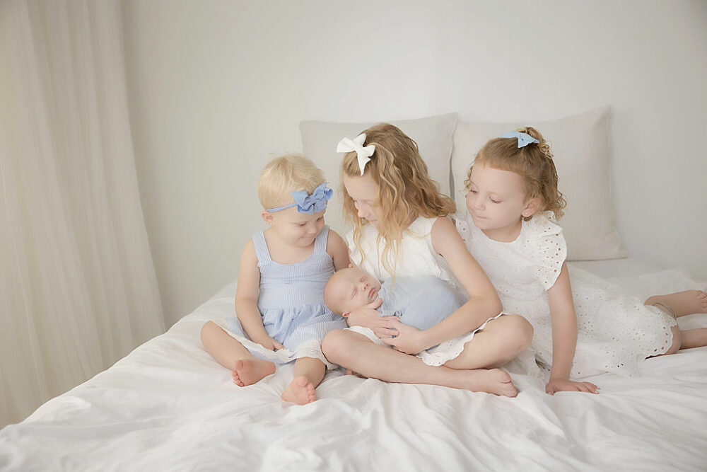 A toddler girl holding her newborn sibling in her arms as he sleeps, sitting next to two sisters on bed photography set for his light-blue newborn session in Eastampton, New Jersey.
