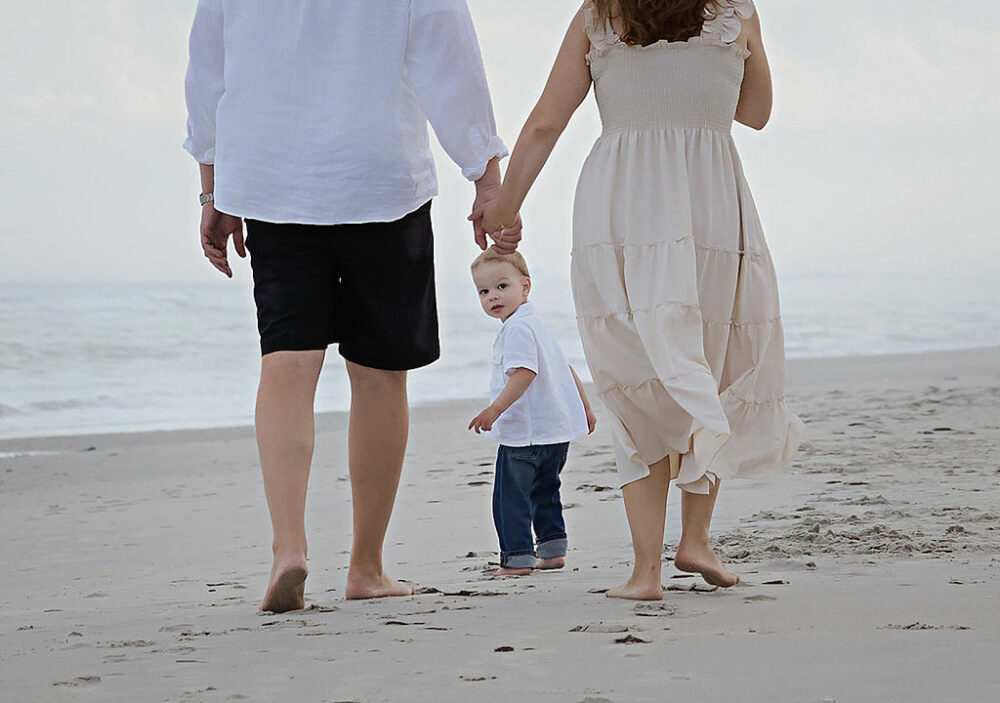 A couple holding hands as their son looks back at them while walking on the beach for the family portrait ideas and summer beach family session in South Jersey.