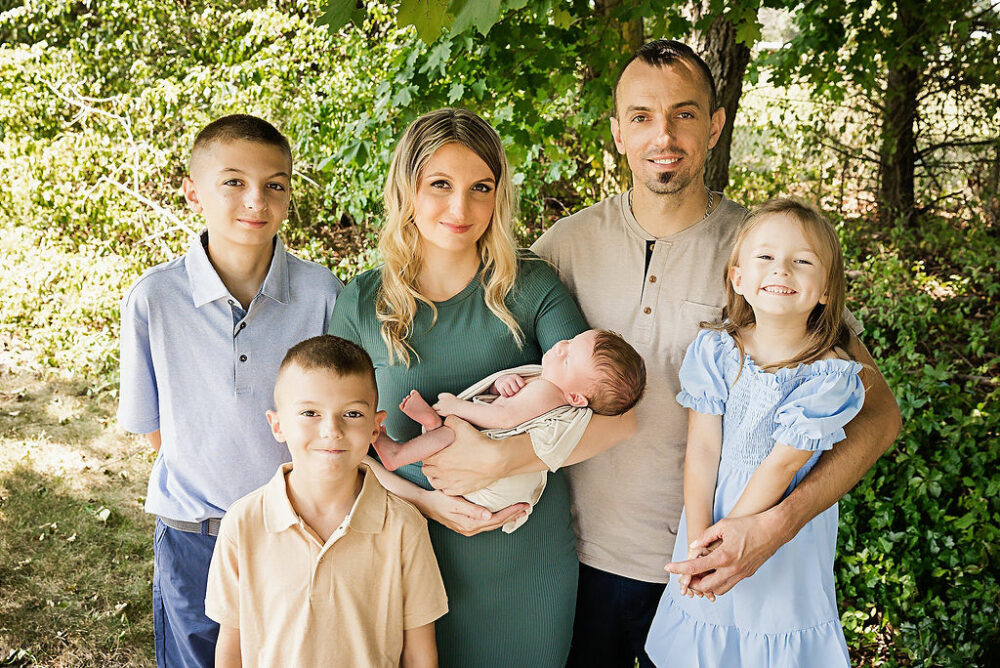 A family photo of man and a woman posing with their four children outdoors in front of greenery wearing neutral colors for their neutral newborn session in Southampton, New Jersey.