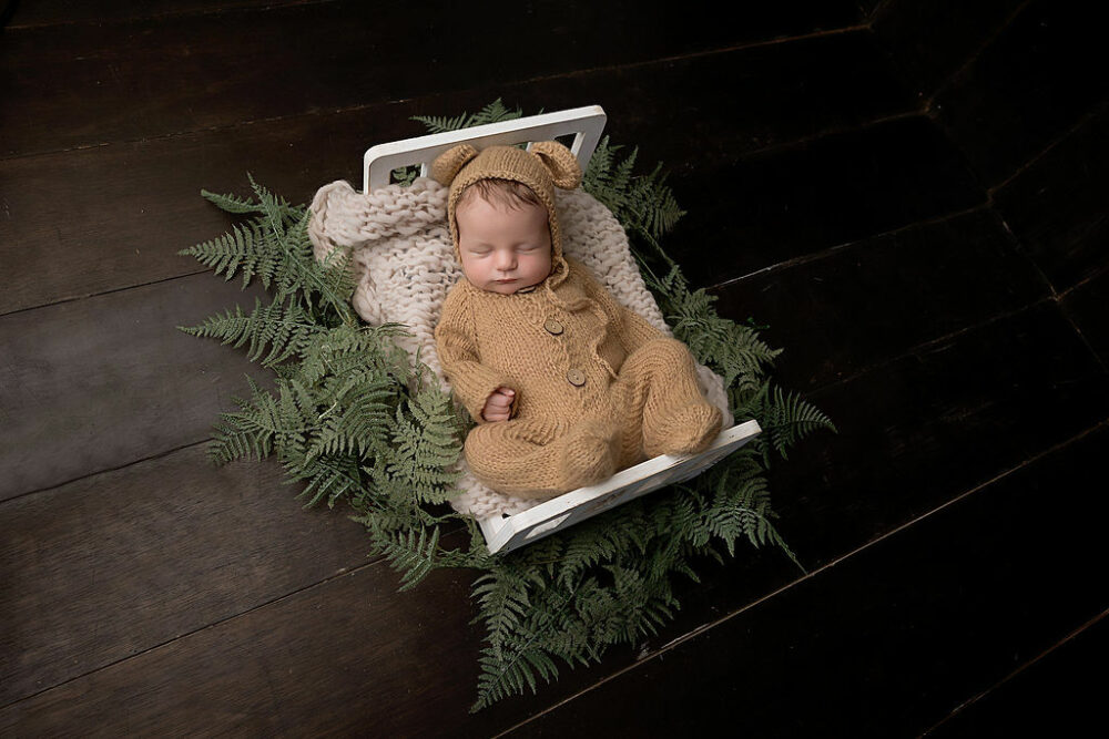 A cute picture of a newborn boy in Bear outfit sleeping on tiny newborn photography crib crib for his baby photoshoot in Eastampton, New Jersey.
