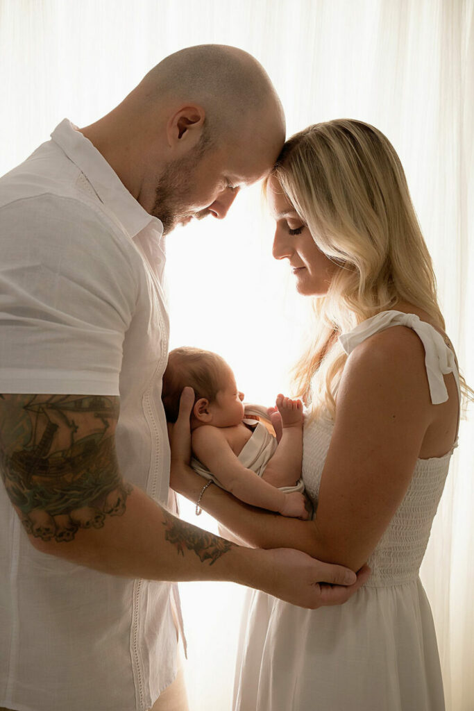 A man and woman facing each other and touching foreheads as she carries their newborn son in her arms close to her chest against a light and bright backdrop for their sons newborn session in Southampton, New Jersey.