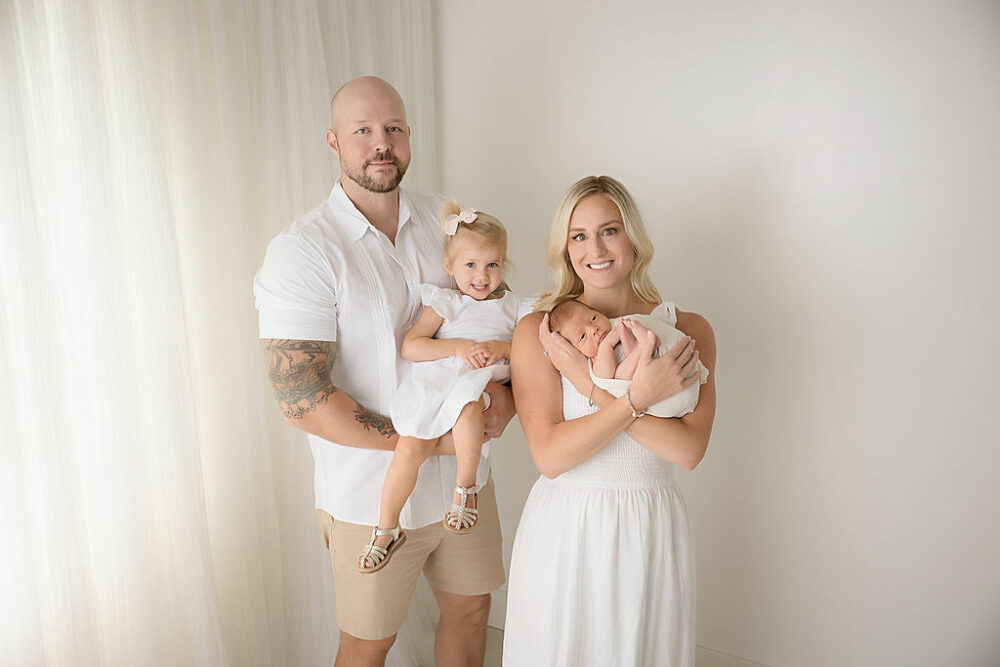 A family portrait of a couple posing and smiling with her two children against a white backdrop, posed for a navy blue newborn session in Westampton, New Jersey.
