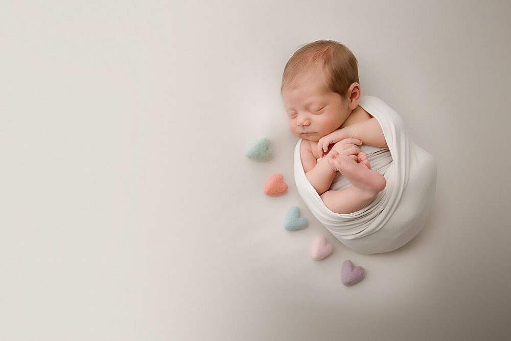 A newborn boy sleeping on his back and swaddled showing his cute feet and hands adorned with heart photography props for his newborn session in Eastampton, New Jersey.