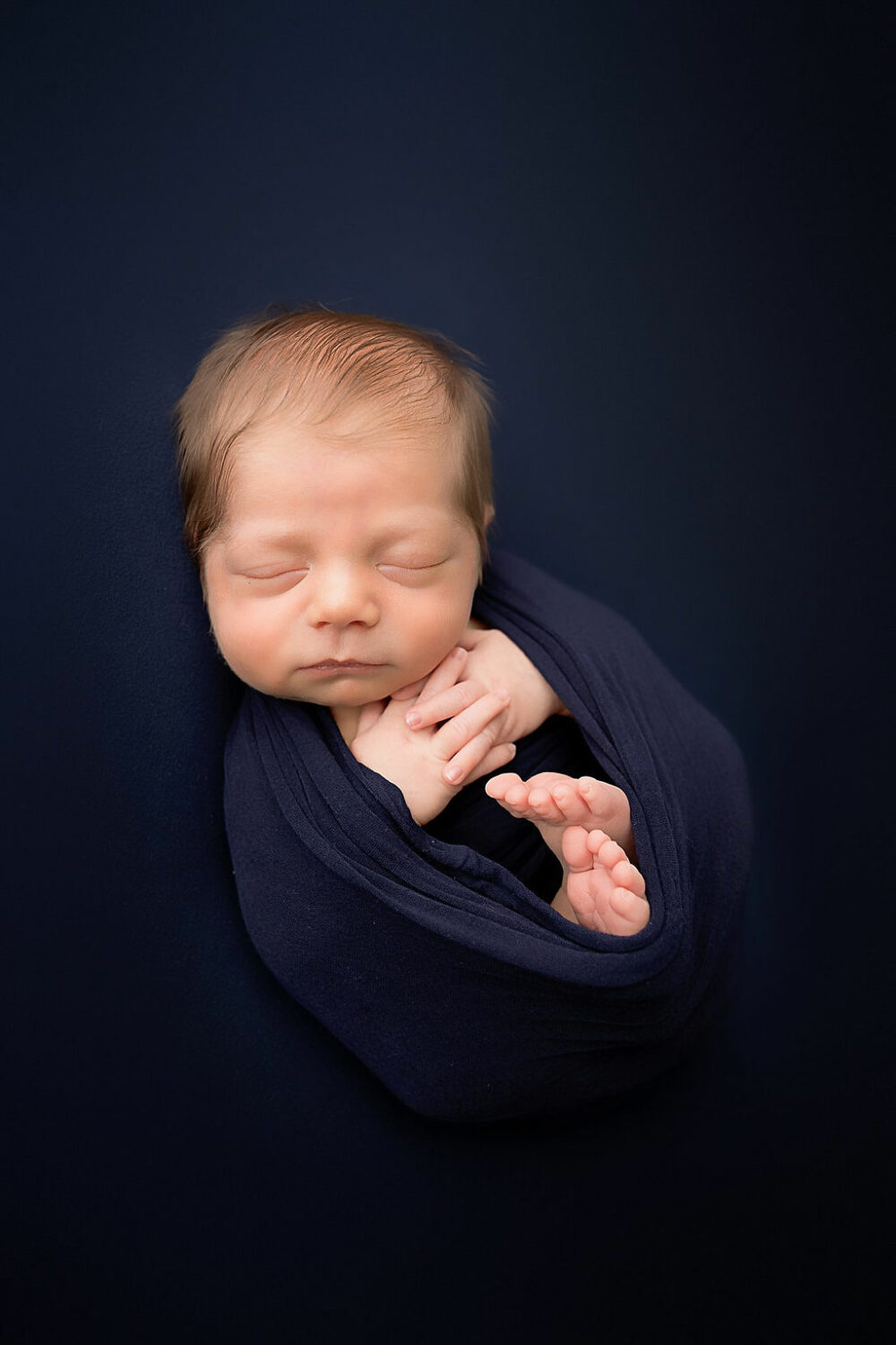 An infant portrait of a sleeping baby boy swaddled but his toes and hand are showing for his professional newborn photography session in Cherry Hill, New Jersey.