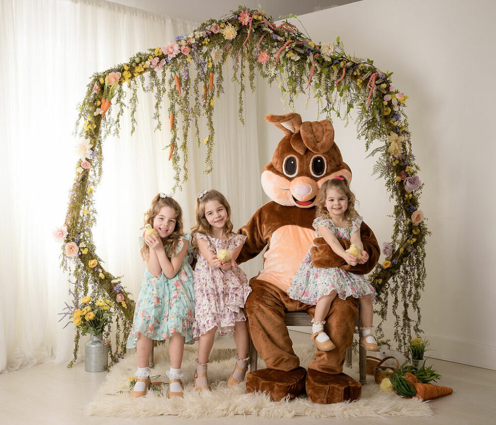 A sibling portrait of three toddler girls sitting on a bench with the Easter bunny adorned with greenery and carrots for there in studio Easter mini session in Camden, New Jersey.