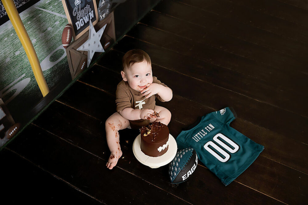 A portrait of a boy eating cake and sitting on the floor next to a jersey and football, looking up during his football first birthday session in Eastampton, New Jersey.