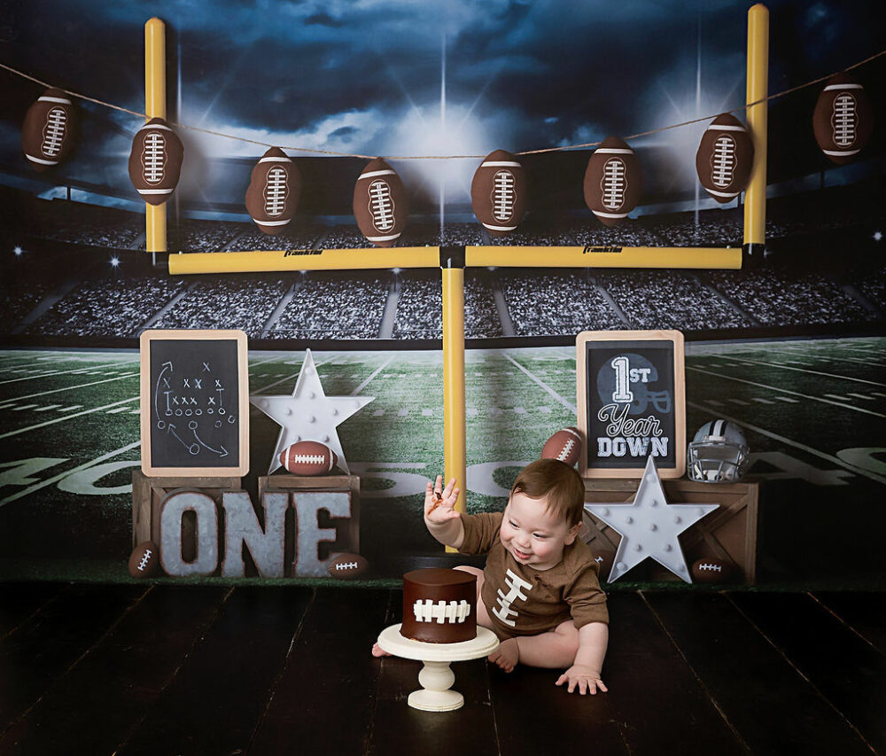 A adorable photo of a boy sitting on the floor with a football stadium theme backdrop with sports theme photography props, eating cake for his Football first birthday session in Westampton, New Jersey.