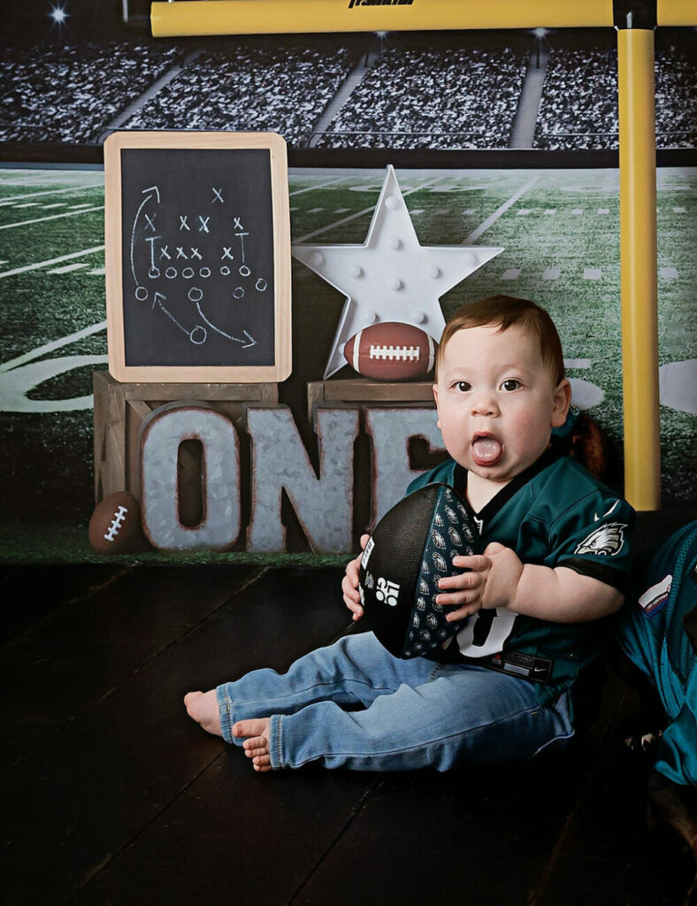 A close-up of a boy sitting and holding a football next to sports themed props for his football first birthday session in Cherry Hill, New Jersey.