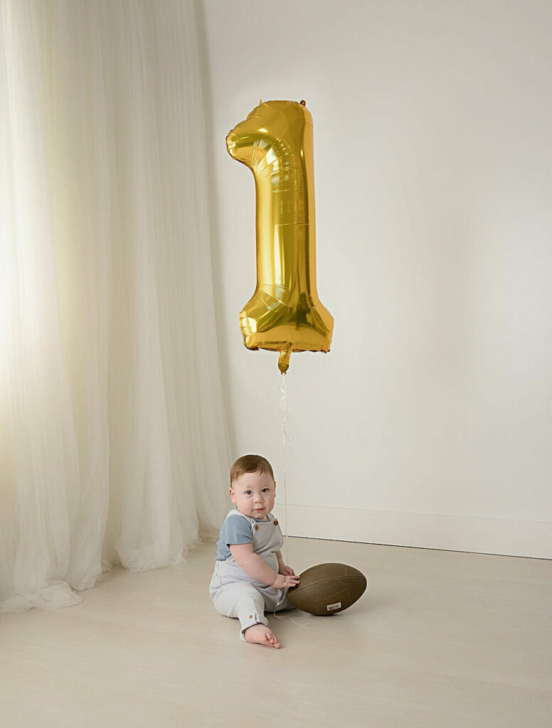 A birthday portrait of a boy sitting on the floor and playing with a football with a balloon number one for his baby Development pictures taken in medford, New Jersey.