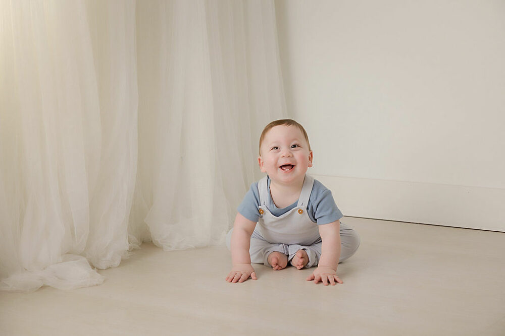 An adorable portrait of a baby boy sitting and smiling at the camera, wearing overalls against a white backdrop for his football first birthday session in Deptford, New Jersey.