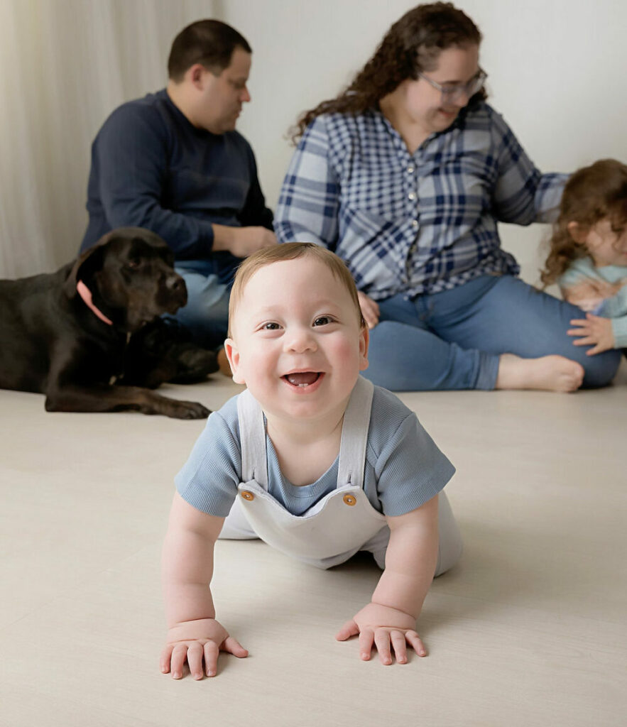 A family photo of a boy crawling and laughing during his first birthday photoshoot with his family in the background during his milestone session in Delran, New Jersey.