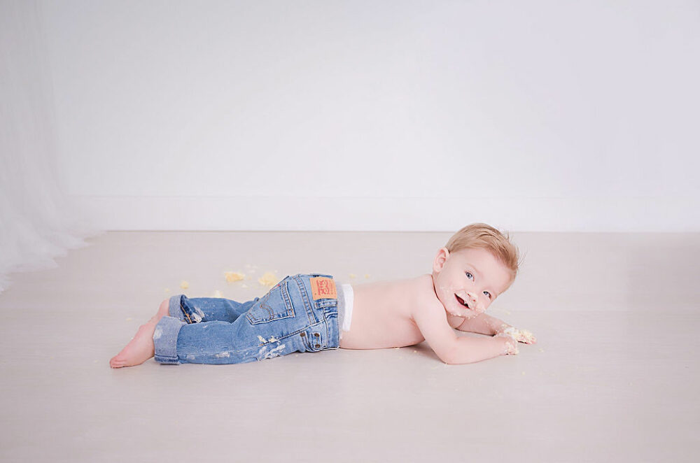 Can you tell her boy, smiling and laying on his belly with a messy face from eating cake and wearing pants for his simple lifestyle first birthday session in Westampton, New Jersey.