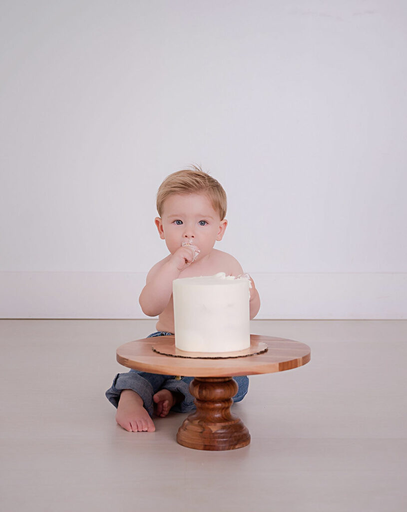 A toddler boy sitting on the ground and eating cake wearing denim jeans for his Simple Lifestyle first birthday session in Southampton, New Jersey.
