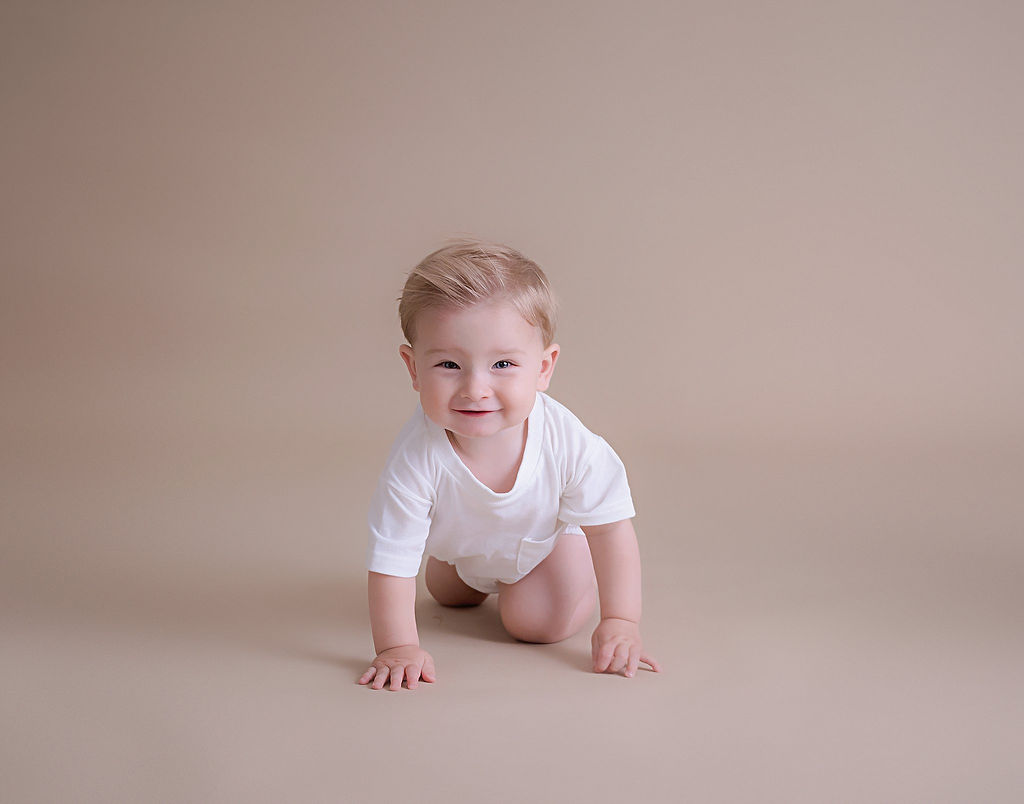 A smiling boy, crawling on the floor, wearing T-shirt for first birthday pics taken during his simple lifestyle first birthday session in Eastampton, New Jersey.