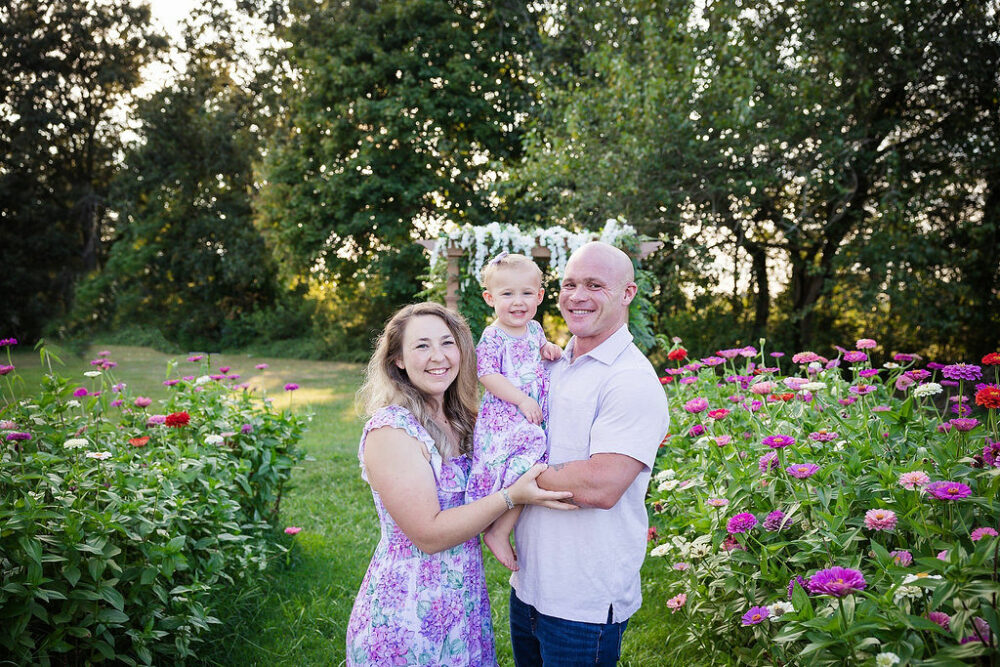 A man and woman standing and carrying their toddler between them and smiling while standing in a flower field for their family flower garden mini sessions in Southampton, New Jersey