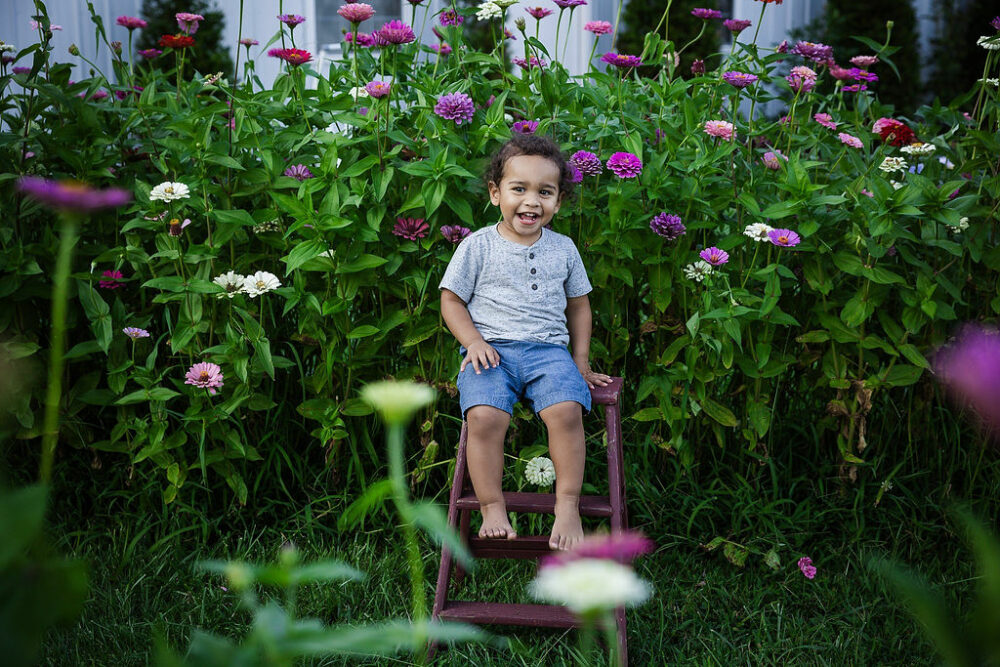 A toddler boy smiling and sitting on a little ladder in a garden for flower minis in Westampton, New Jersey.